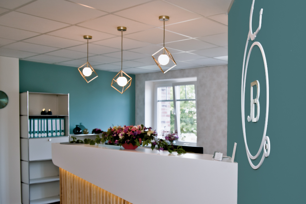 Modern eingerichteter Empfangsbereich einer Praxis mit einem weißen Tresen, der teilweise mit Holzlamellen verkleidet ist. Im Hintergrund befinden sich ein Regal, ein Computer und zwei große Fenster, die viel natürliches Licht hereinlassen. Die Wände sind in einem beruhigenden Blau- und Grauton gestrichen, und drei stilvolle Hängeleuchten hängen von der Decke.