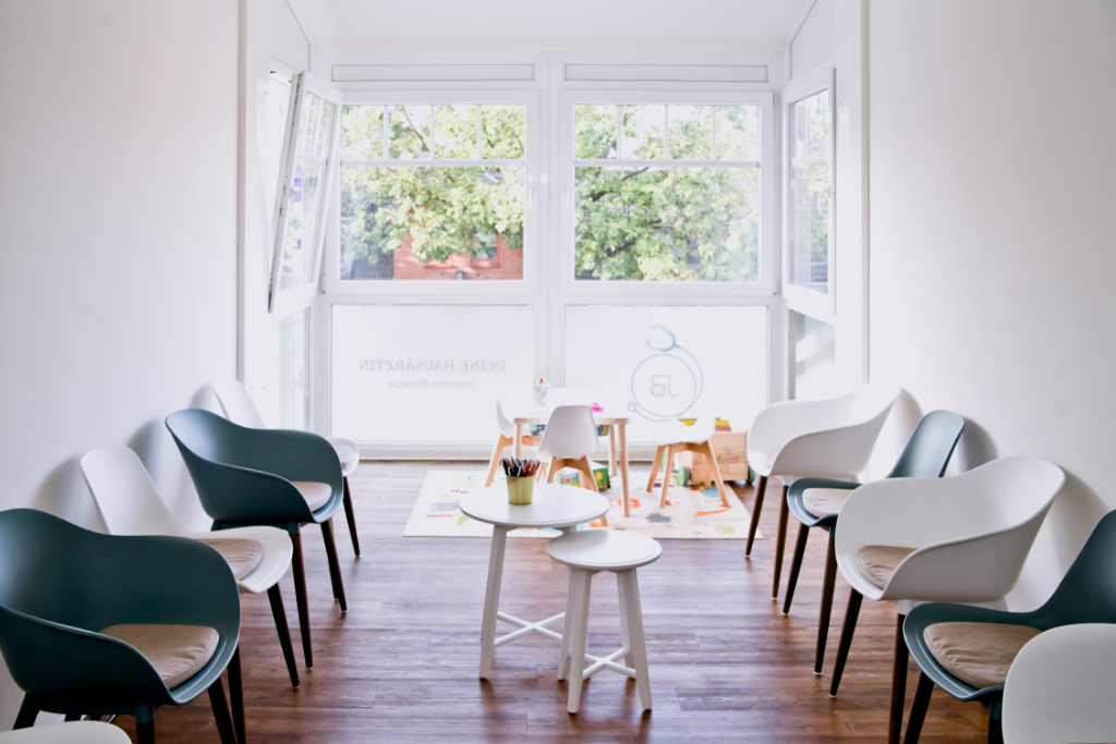 Modern eingerichteter Empfangsbereich einer Praxis mit einem weißen Tresen, der teilweise mit Holzlamellen verkleidet ist. Im Hintergrund befinden sich ein Regal, ein Computer und zwei große Fenster, die viel natürliches Licht hereinlassen. Die Wände sind in einem beruhigenden Blau- und Grauton gestrichen, und drei stilvolle Hängeleuchten hängen von der Decke.